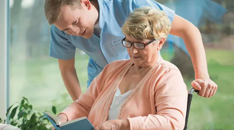 Pflegehilfe unterstützt in den eigenen vier Wänden eine Frau im Rollstuhl.